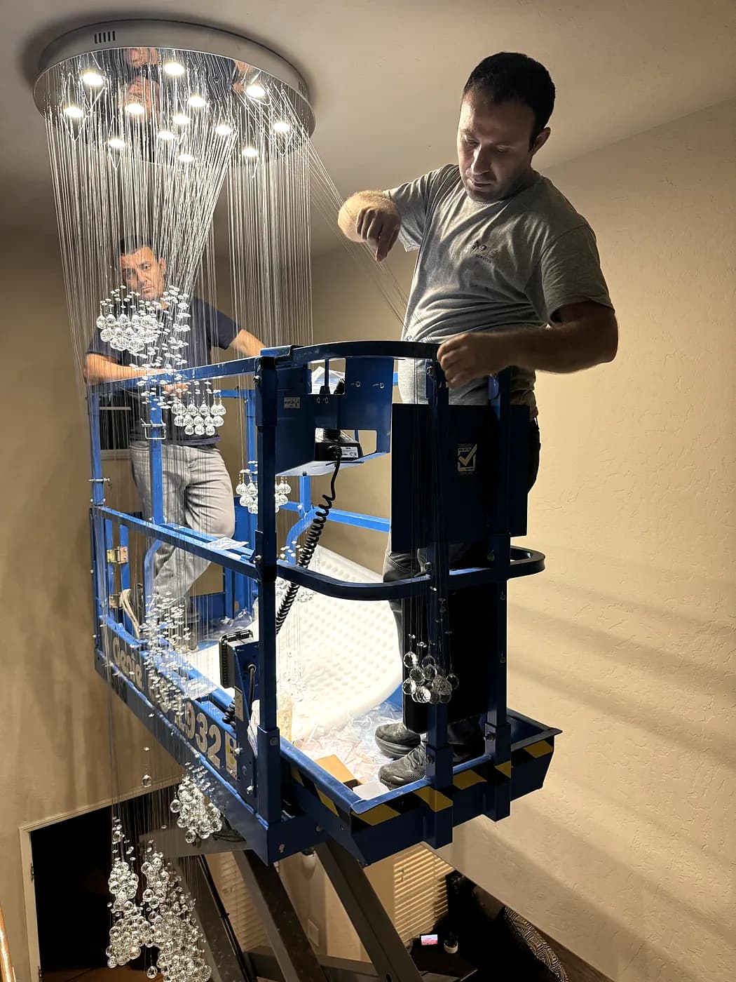 A ProLine electrician on a lift installing a large chandelier under a high ceiling